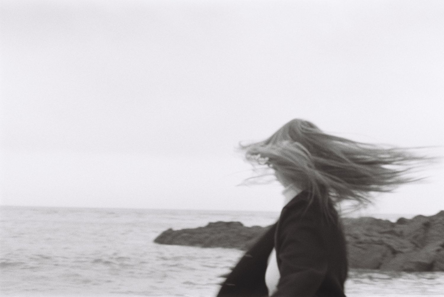 Dramatic black and white photo of wind-blown hair against rocky coastal landscape.