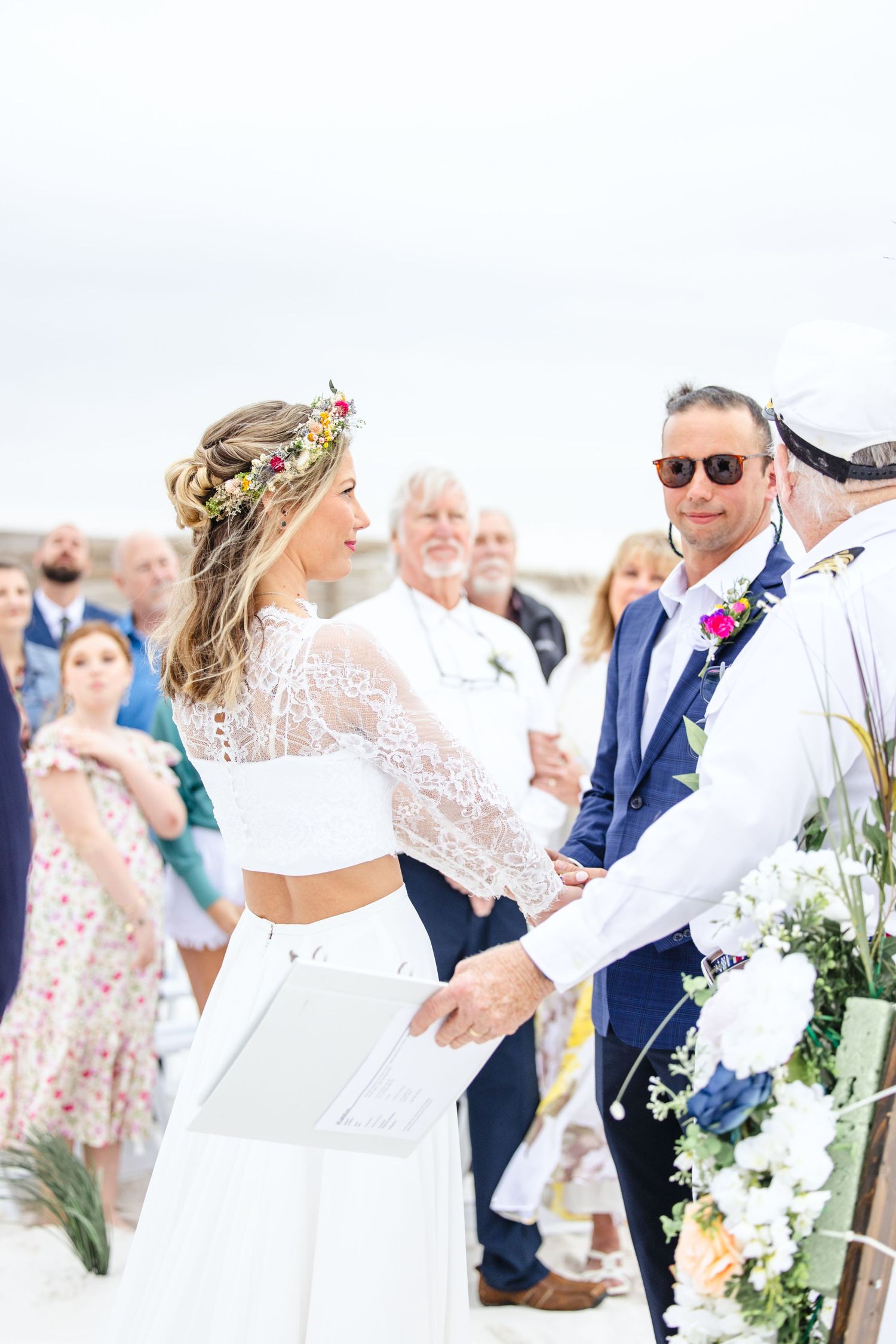 Beach wedding ceremony with guests watching couple exchange vows under floral decorations.