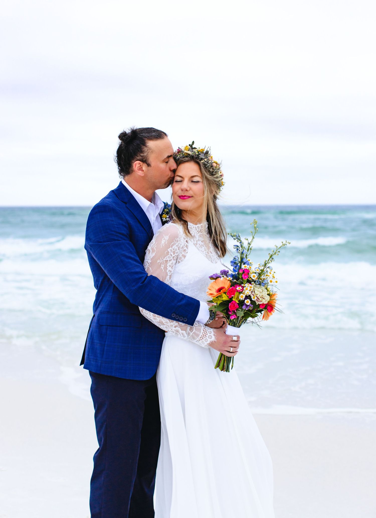 A romantic beach wedding moment with a couple embracing in formal attire and floral bouquet against ocean waves.