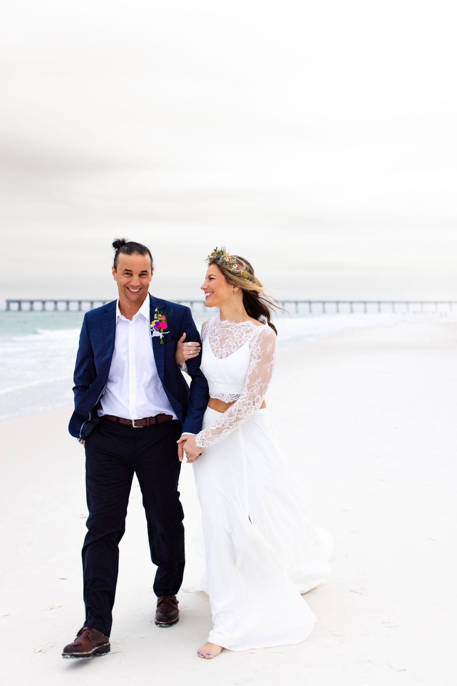 A couple walking barefoot on white sand beach with pier in background, wearing wedding attire.