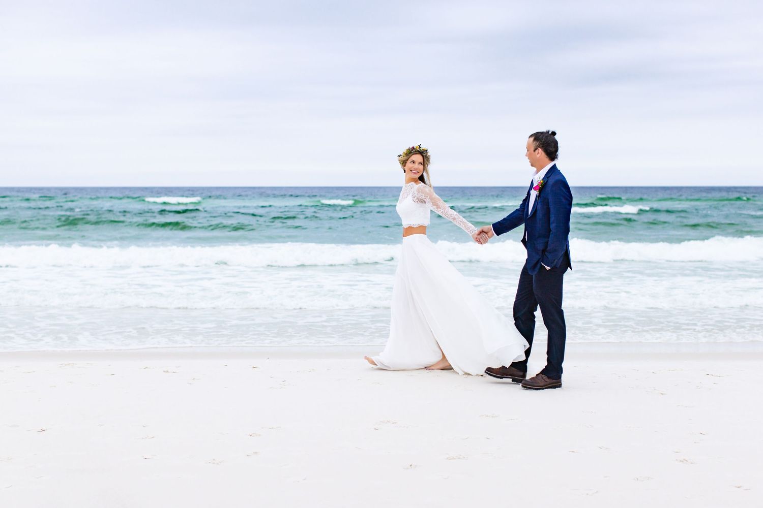A couple in wedding attire holds hands while dancing on a pristine white sand beach.