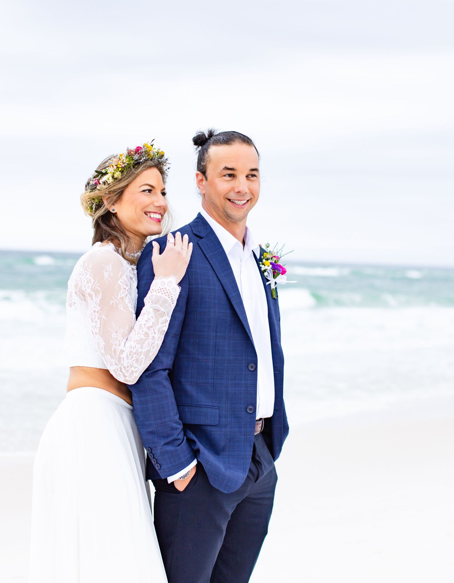 A happy couple poses on beach in wedding attire with flower crown and bright smiles.