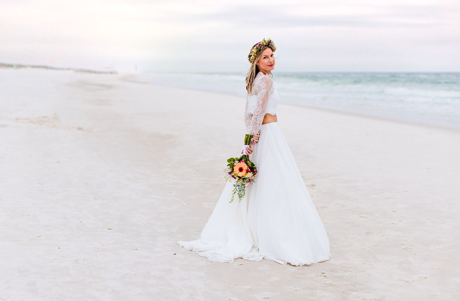 A bride in lace dress holds colorful bouquet while walking alone on pristine beach.