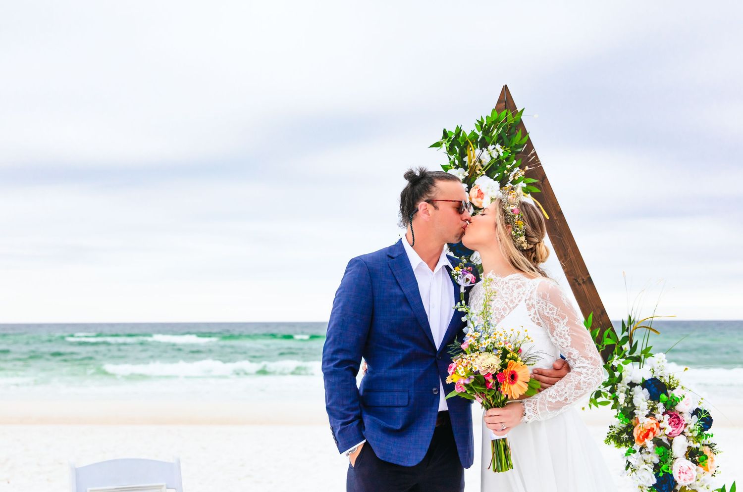 Beach wedding ceremony with couple kissing under triangular floral arch by ocean.