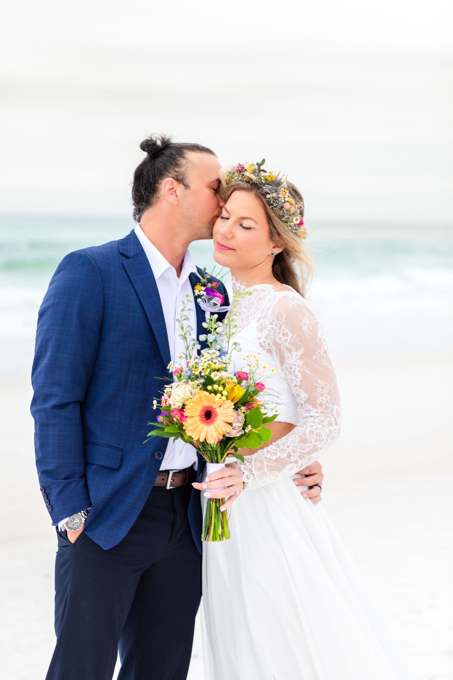 Newlyweds share intimate moment on beach, bride holding colorful sunflower bouquet.