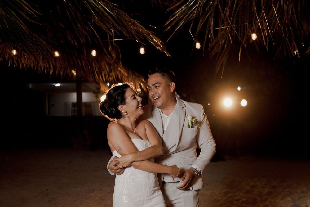 A couple shares their first dance under a thatched roof with warm string lights at a tropical beach wedding.