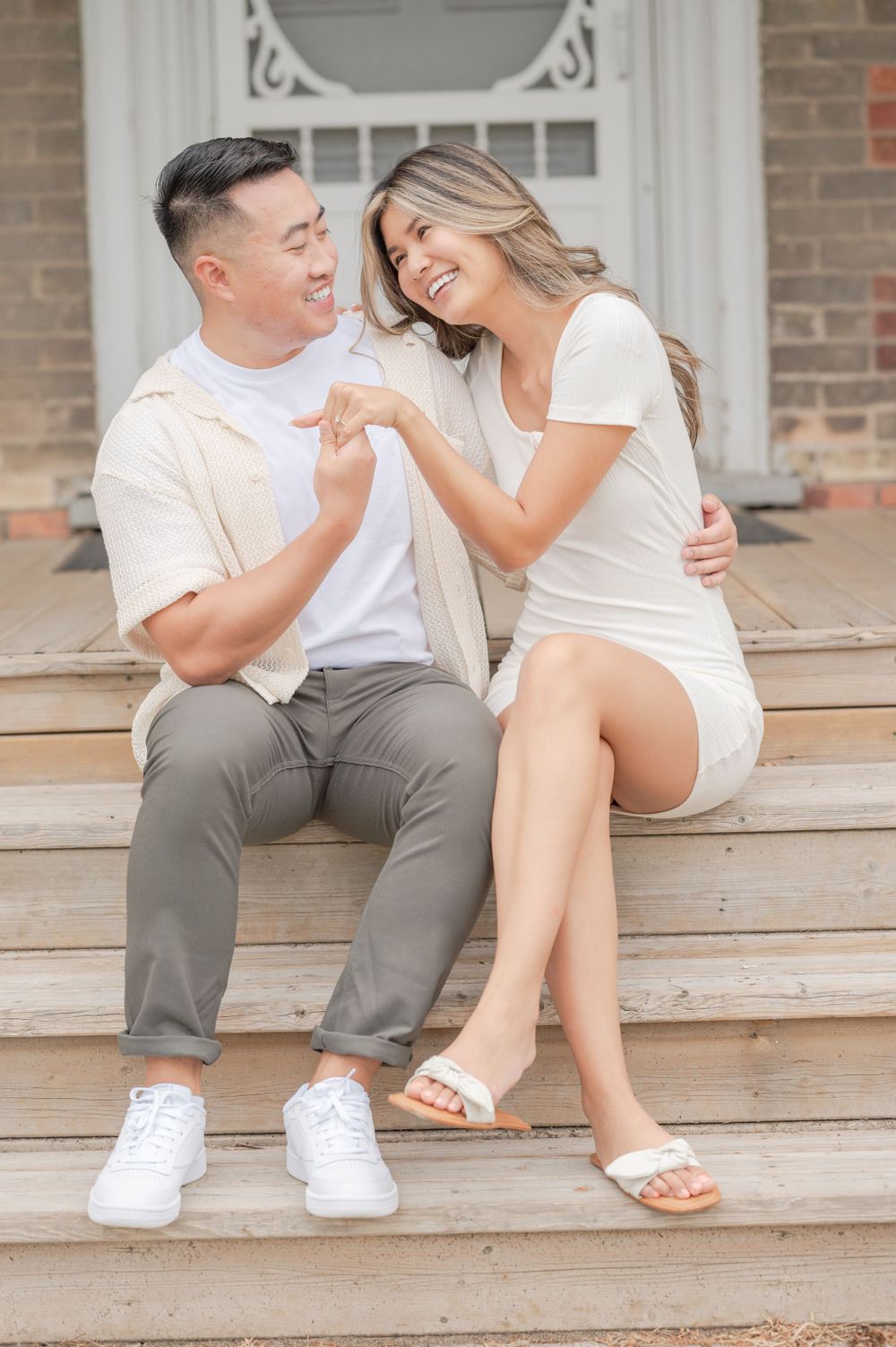 A couple in white and grey outfits sitting on wooden steps outside a brick house with a white door.