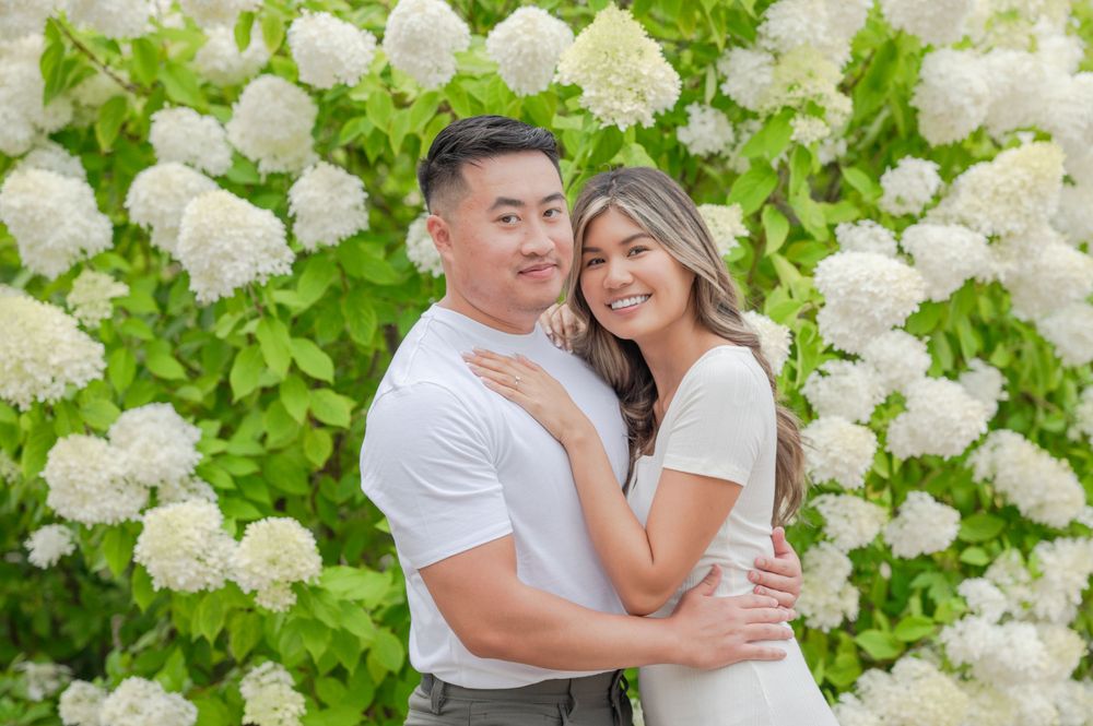 A couple poses together against a backdrop of blooming white hydrangea flowers.