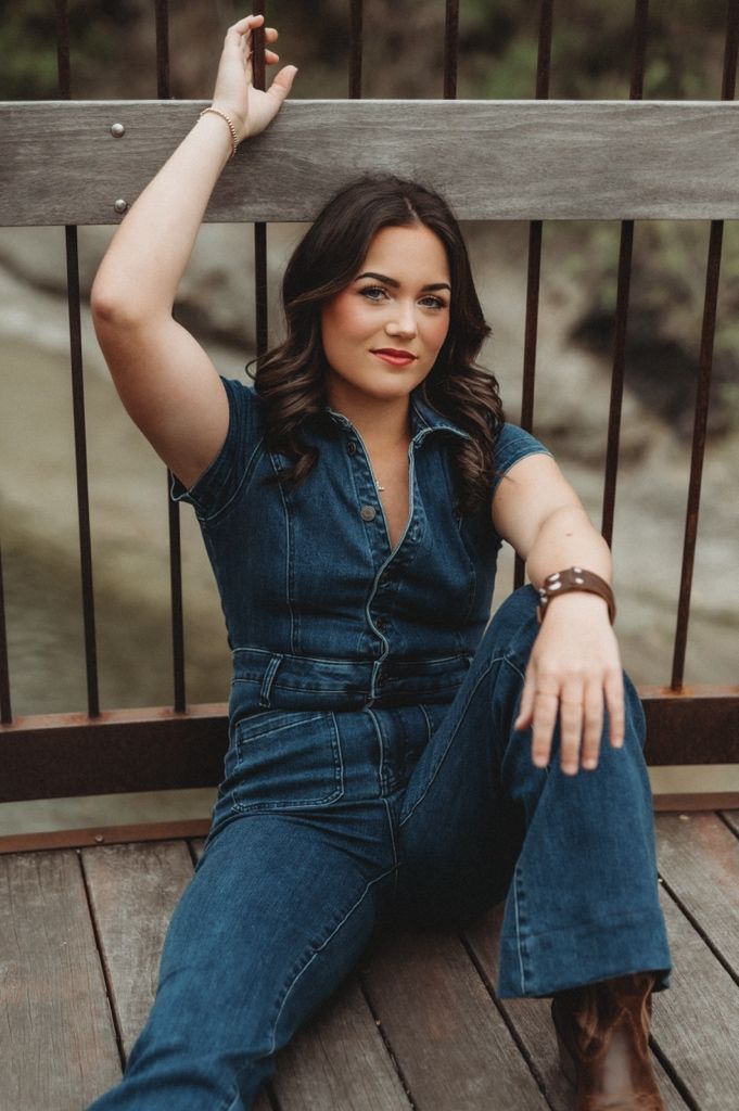 Woman in denim jumpsuit and boots poses casually on wooden deck.