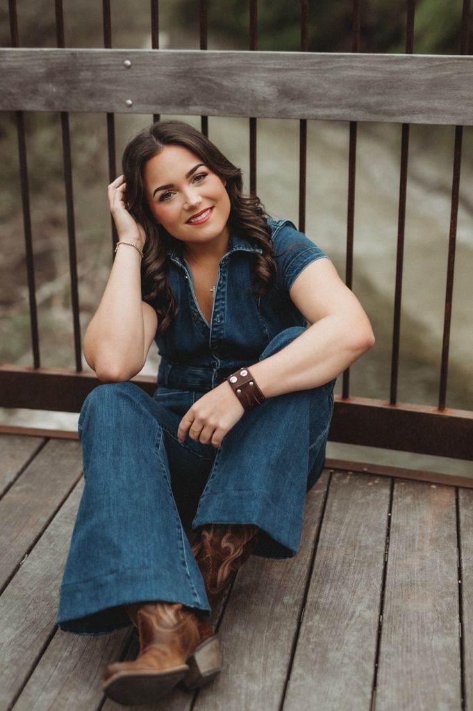 Woman in denim outfit sits relaxed on wooden deck wearing boots.