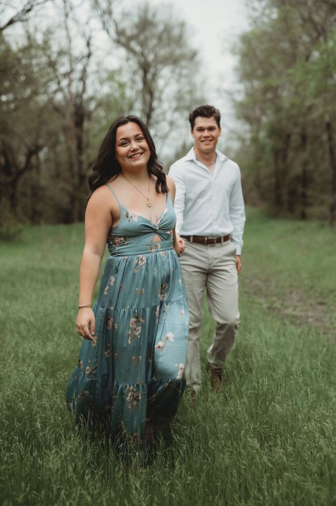 A couple poses in a grassy field, with the woman wearing a blue floral dress.