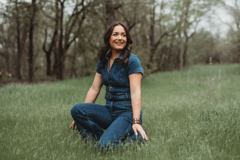 A smiling woman in denim sits in a grassy field during a portrait session.