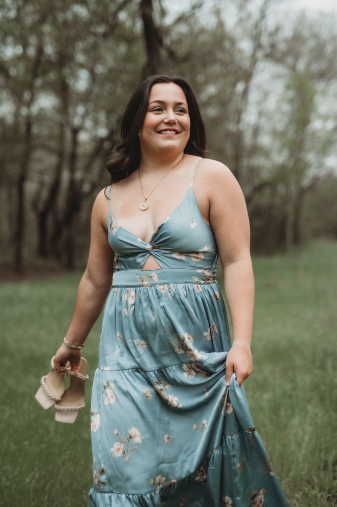 Woman in a blue floral dress holds shoes while posing in a grassy outdoor setting.