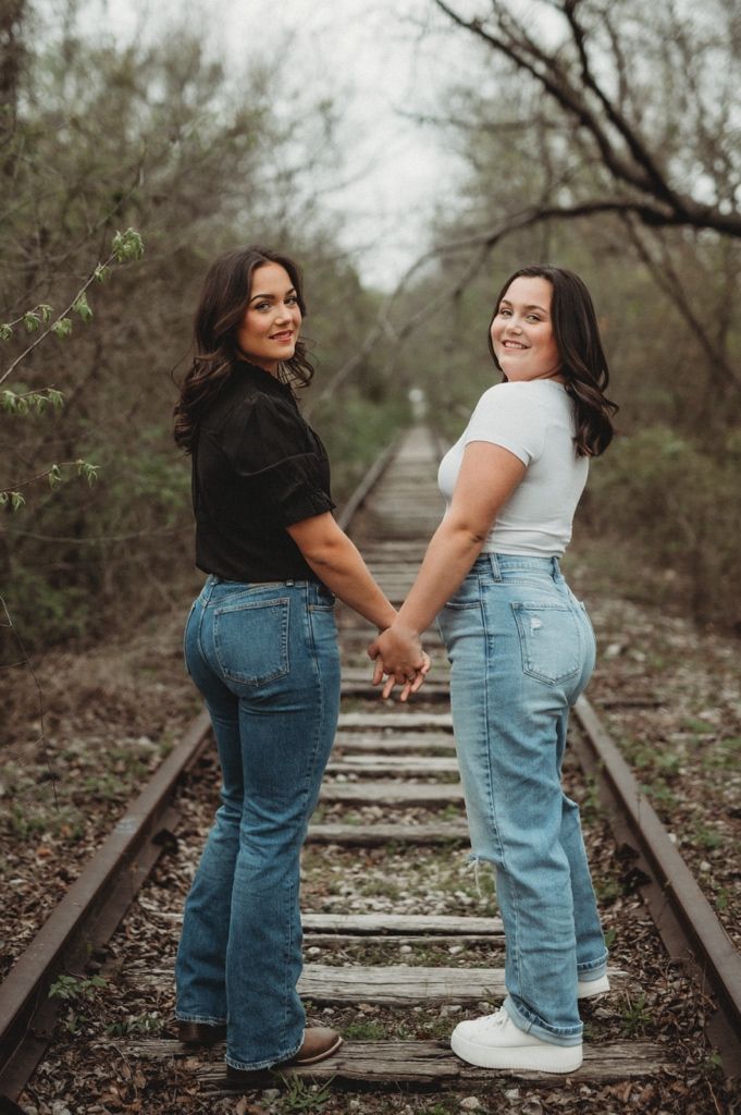 Two people hold hands while standing on railroad tracks in casual denim outfits.