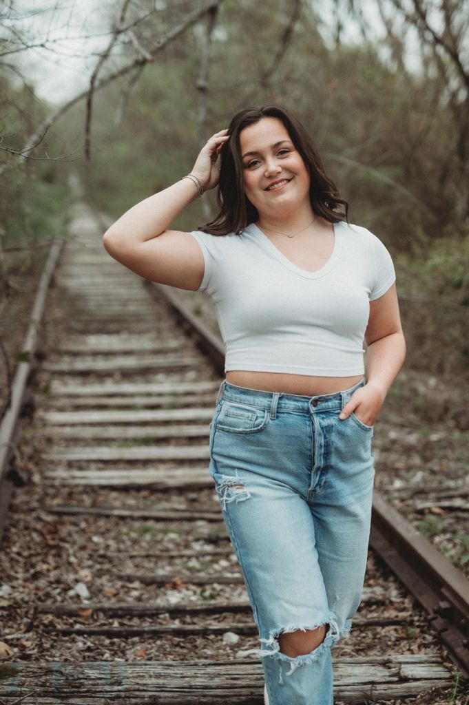 Woman in white crop top and ripped jeans poses on railroad tracks with a bright smile.