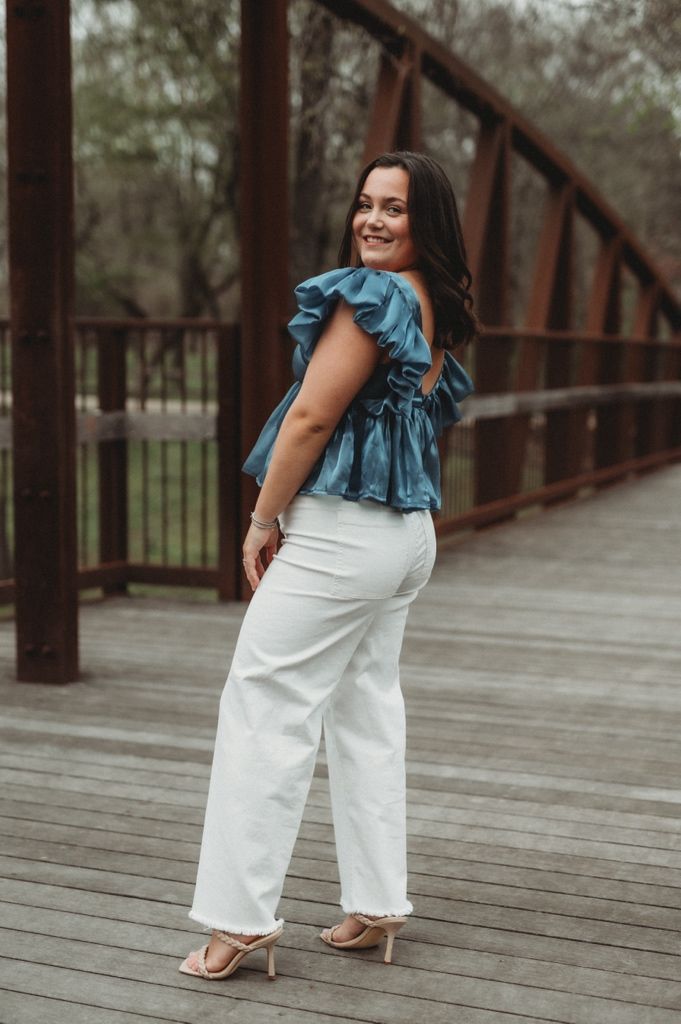 Woman in stylish blue top and white pants poses on wooden bridge.