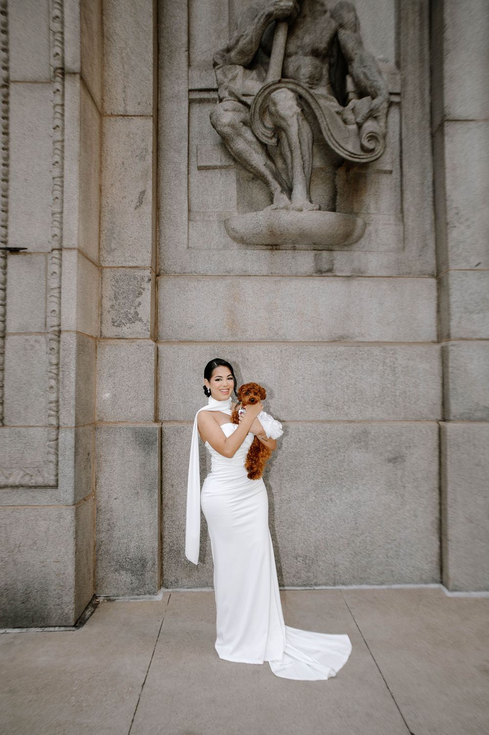 A bride in a flowing white gown sits against a stone wall holding a brown teddy bear in an artistic photo series.