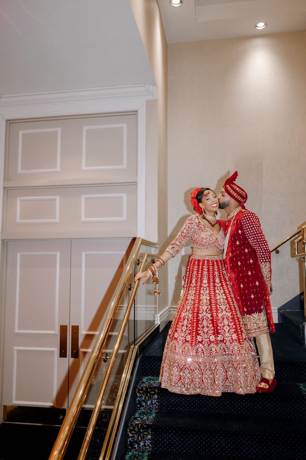 Indian bride and groom in red traditional wedding attire share a romantic moment on elegant staircase with gold railings.