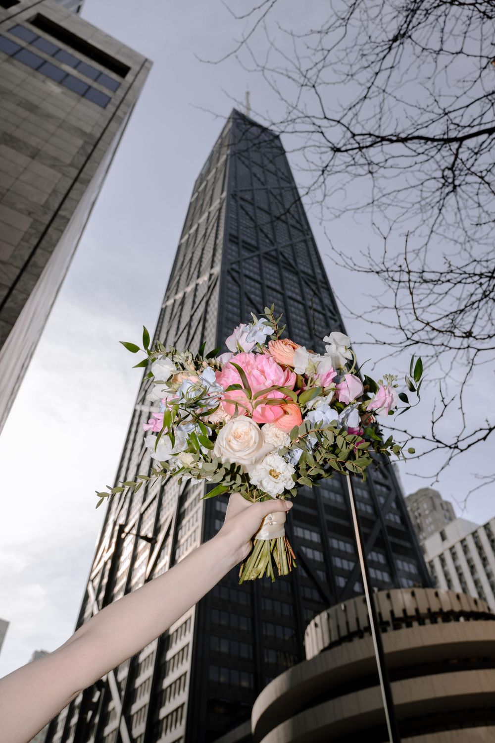 A bouquet of pink and white flowers held up against a modern city skyscraper with bare tree branches in winter.