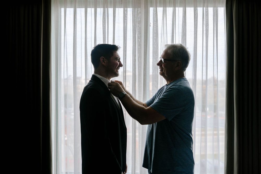 Two silhouettes stand in conversation against a bright window with vertical blinds.