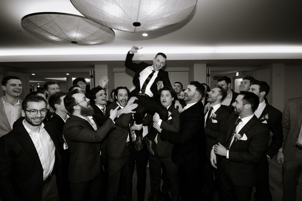 Groomsmen joyfully toss groom into the air during wedding celebration in black and white photo.
