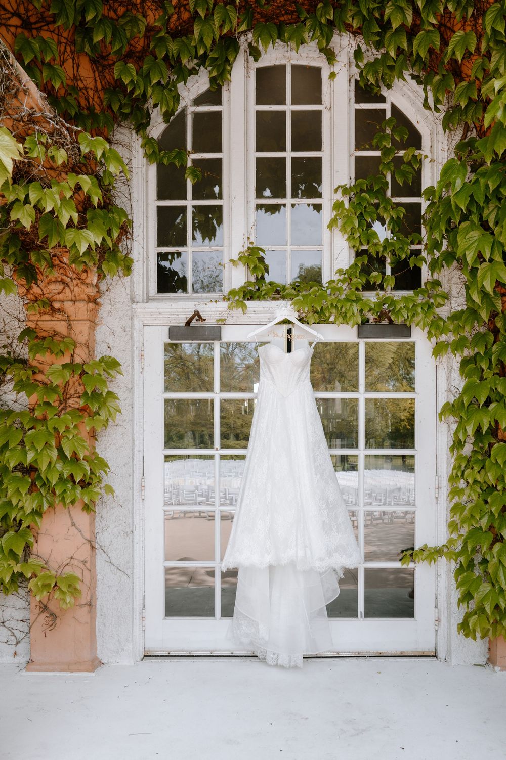 Elegant white lace wedding dress hanging on French doors framed by lush green ivy at wedding venue.