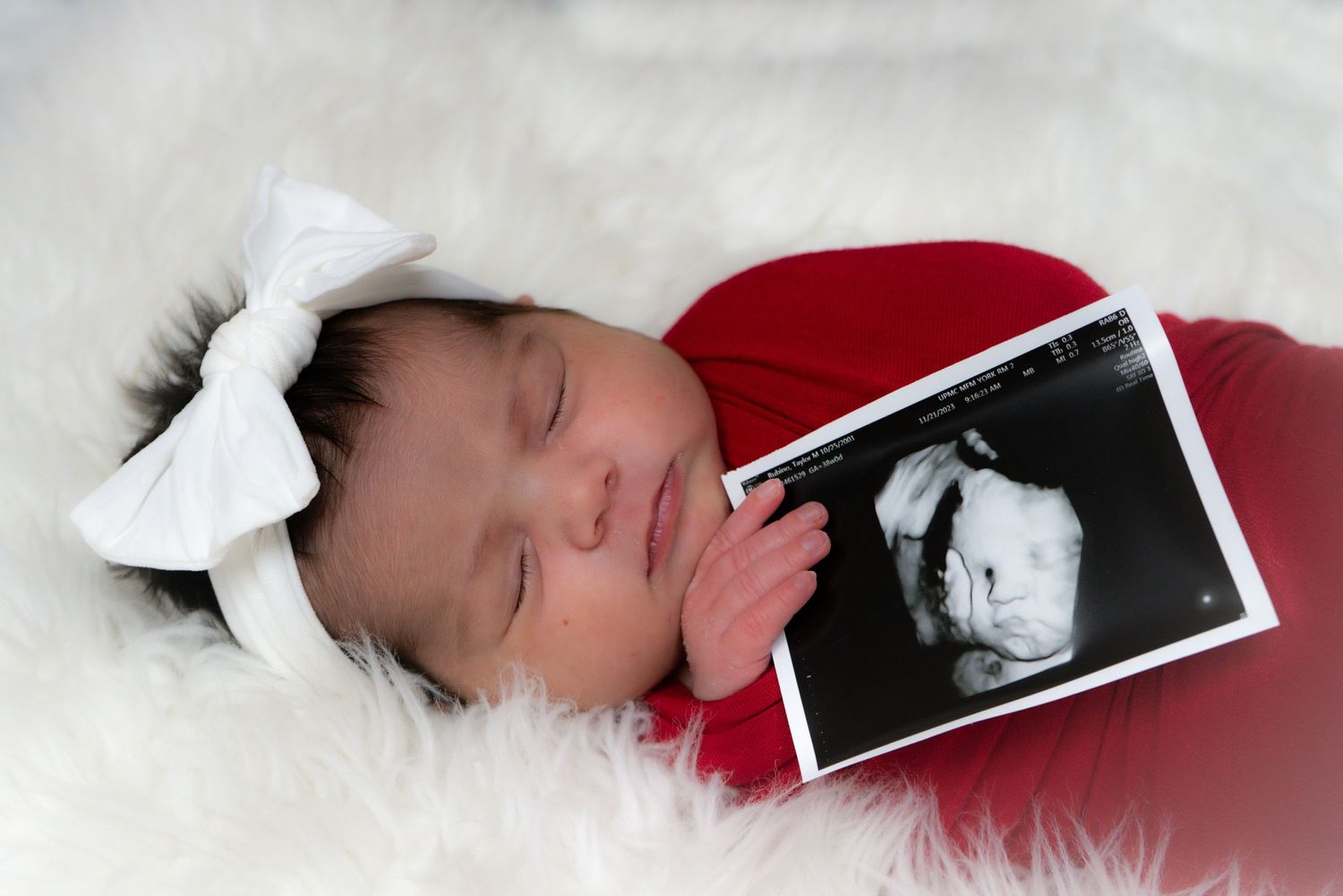 Sleeping baby in red outfit with white bow holding ultrasound photo on white fur backdrop.