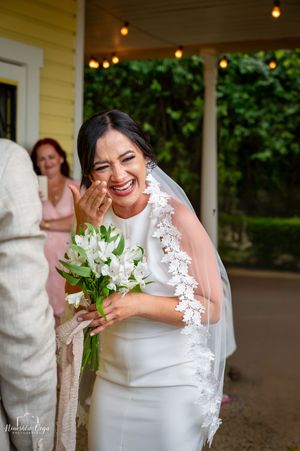 A bride in a white lace dress laughs while holding a bouquet of white lilies on a covered porch.