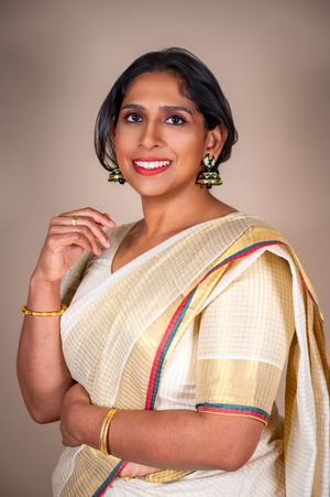 A person wearing a cream-colored traditional Indian saree with colorful border trim poses against a neutral backdrop.