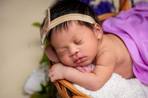 Sleeping newborn wearing a beige headband rests peacefully in a wooden basket with purple fabric.