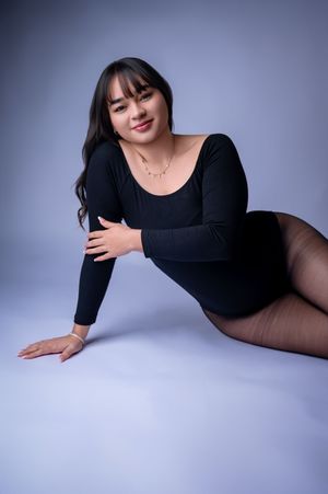 Studio portrait of a person in a black long-sleeve bodysuit posing on a light gray backdrop.