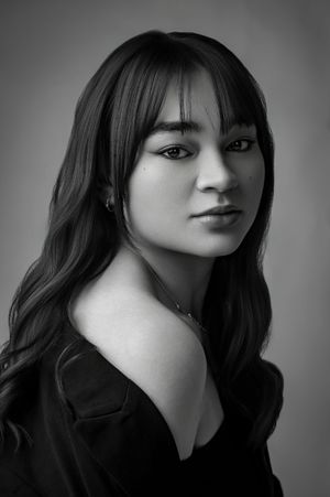 Black and white portrait of a young woman with dark wavy hair and bangs in a black off-shoulder top against gray background.