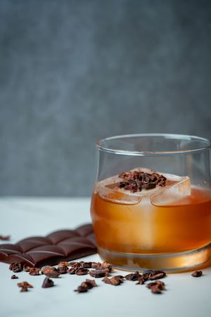 A craft cocktail with bourbon and ice garnished with cacao nibs sits next to a chocolate bar on a white surface.