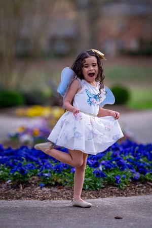 Young child in white dress twirling and jumping by garden of blue flowers.