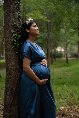 A person in a navy blue dress poses in a serene outdoor setting with trees and greenery in the background.