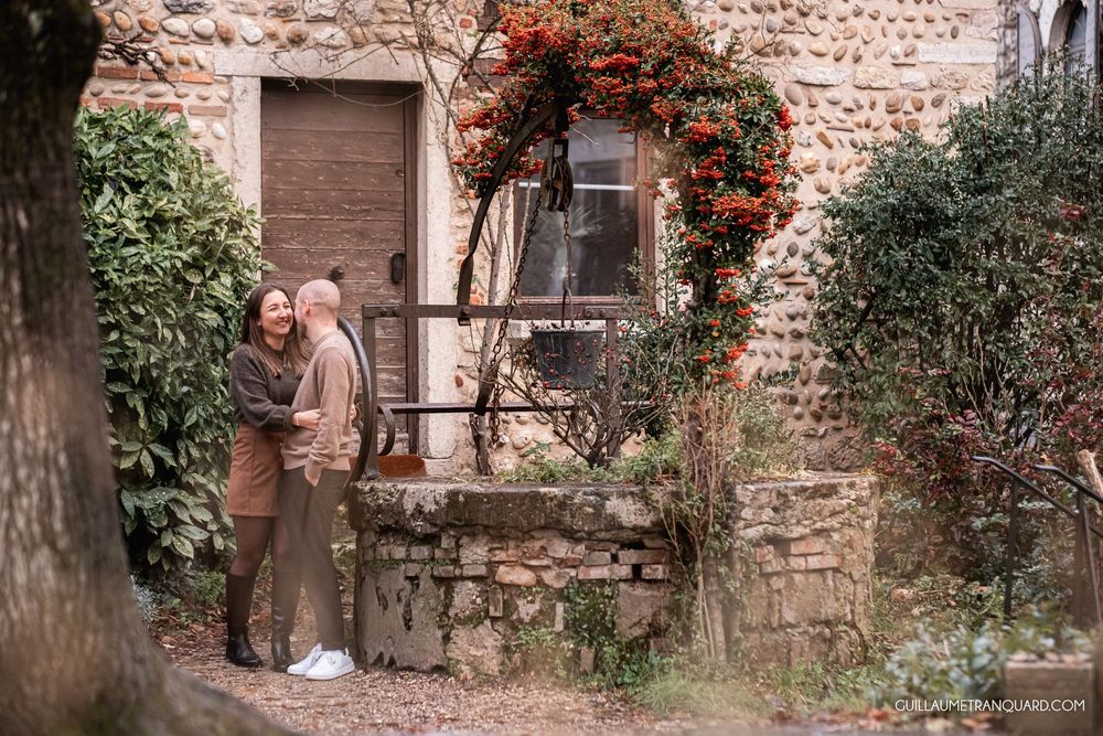 Romantic couple poses against rustic stone wall with climbing red flowers in autumn setting.