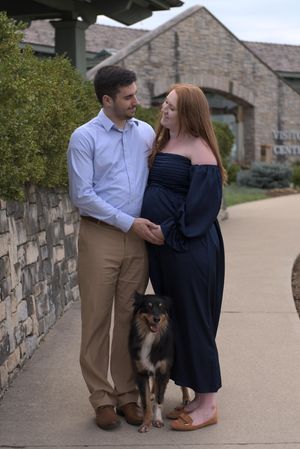 A couple embraces for maternity photos. Their dog stands between them and looks at the camera as the couple looks lovingly at each other.