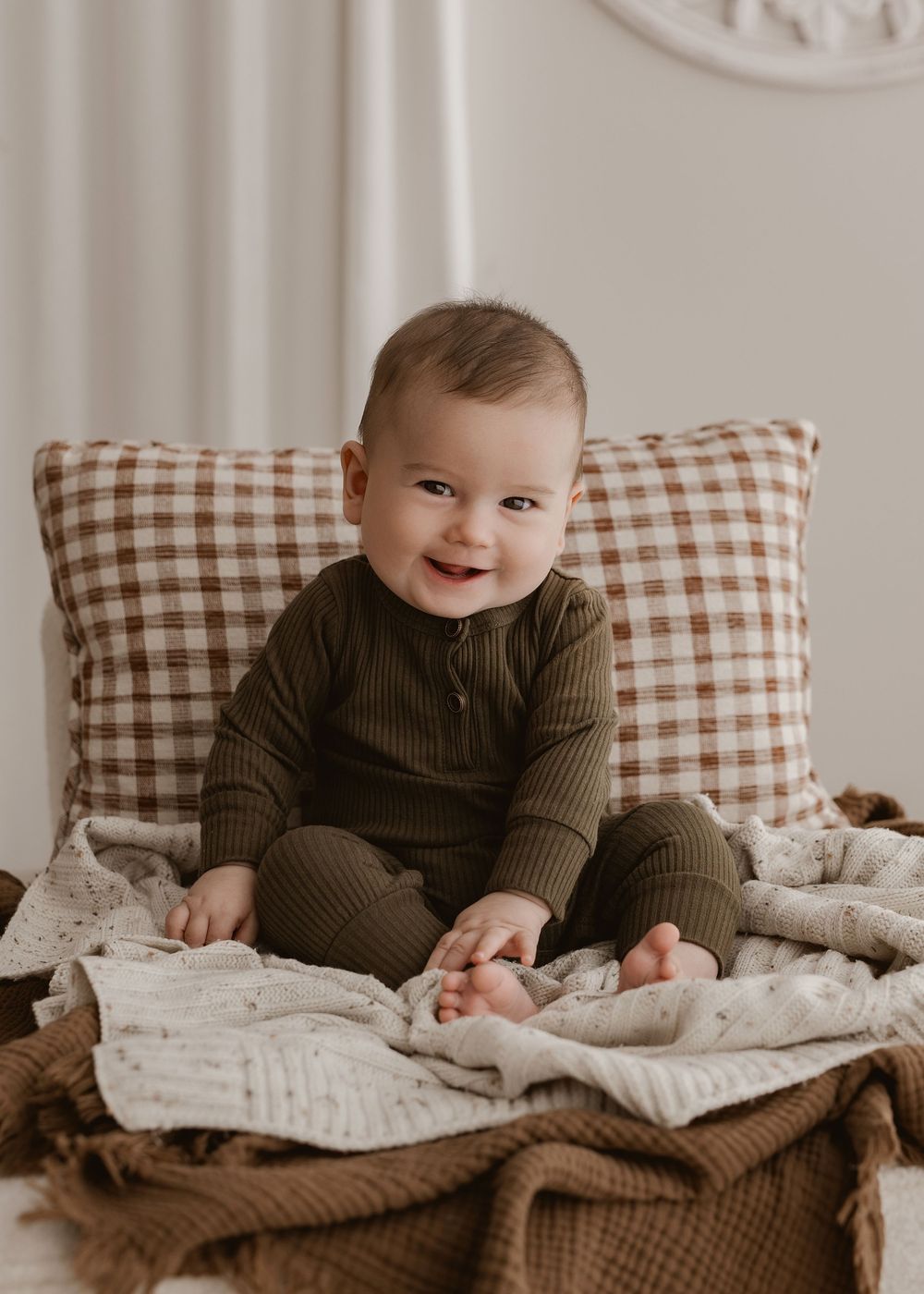 Baby in brown sweater sits on bed with gingham pillows and knit blankets showing progression of happy expressions.