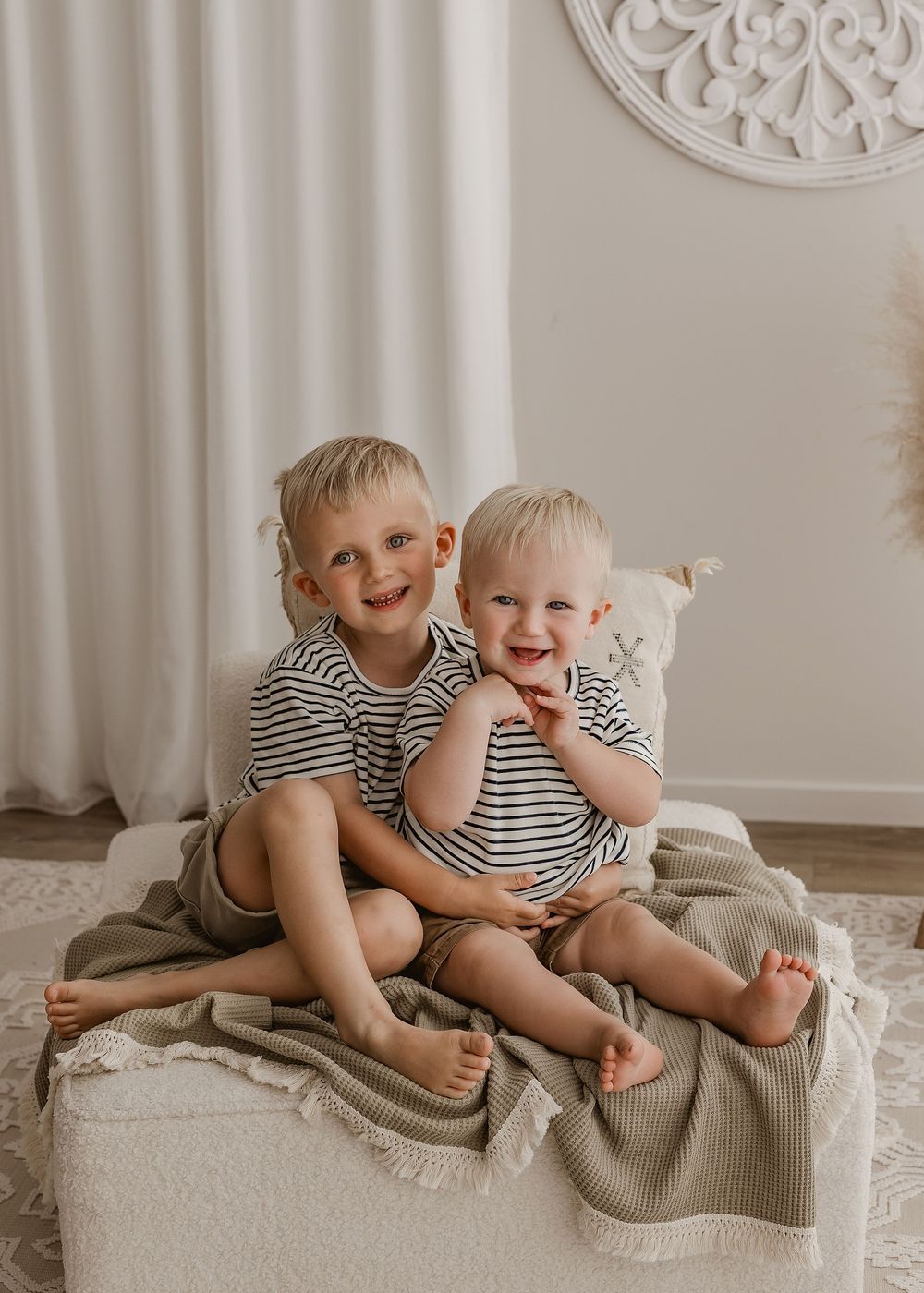 Two children in striped shirts cuddle together on a beige couch in a light-filled room with ornate wall decor.