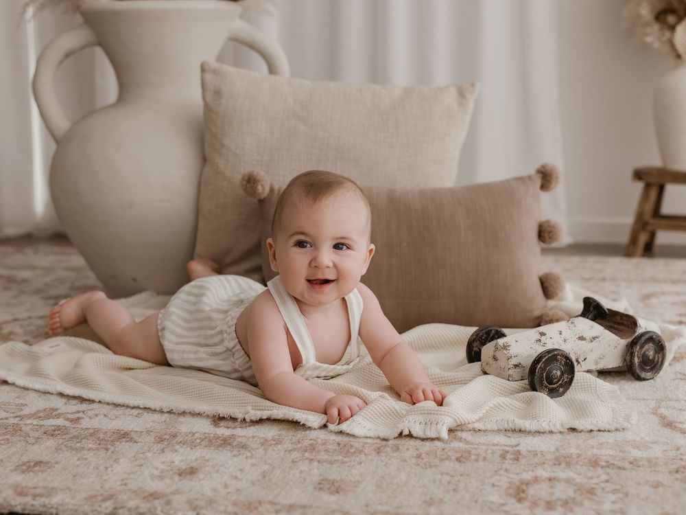 A baby in white clothing lies on a beige carpet while playing with a vintage toy car in a neutral-toned room.