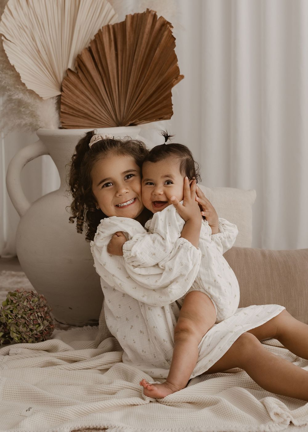 Two figures in white dresses share playful moments on a light-colored bed with palm leaf decor in the background.