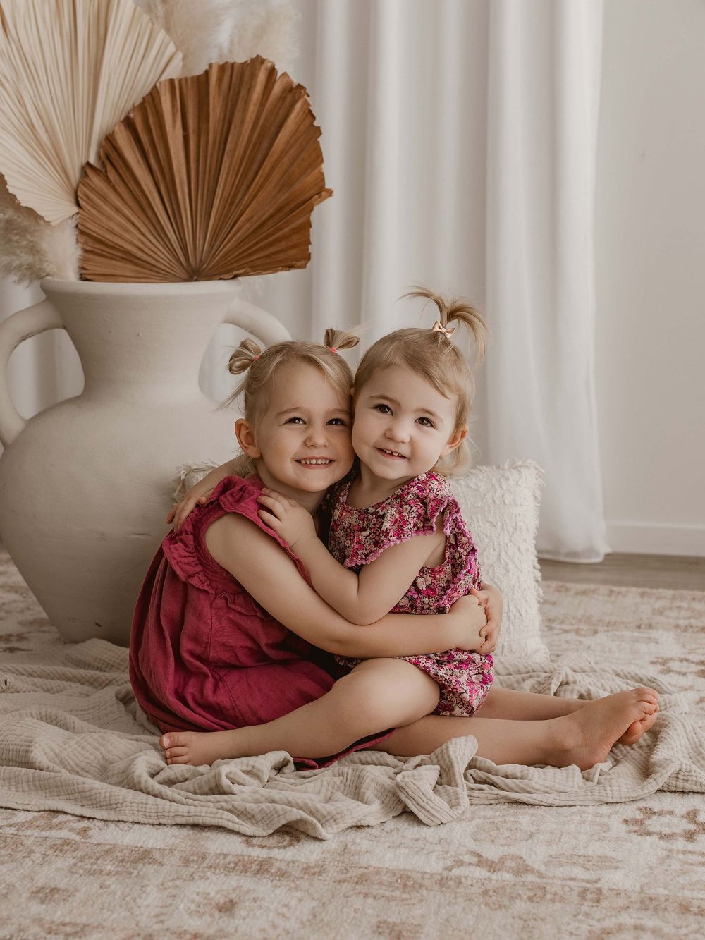 Two children in burgundy and floral outfits share sweet embraces while sitting on a cream-colored floor.