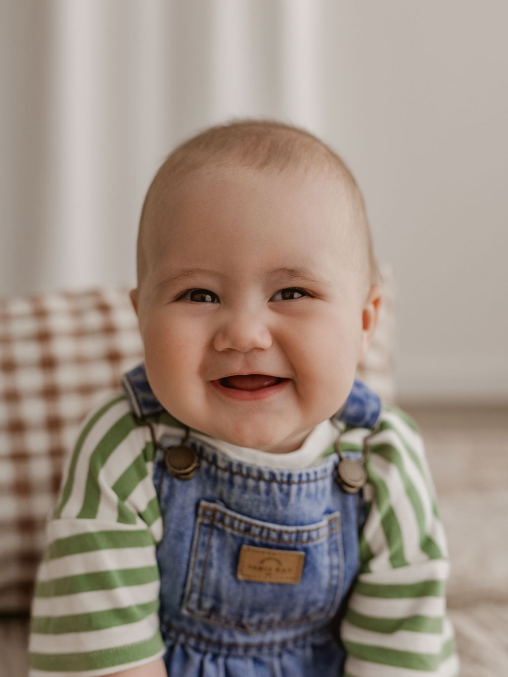 A happy baby wearing denim overalls and a green striped shirt smiles brightly while sitting in front of a plaid chair.