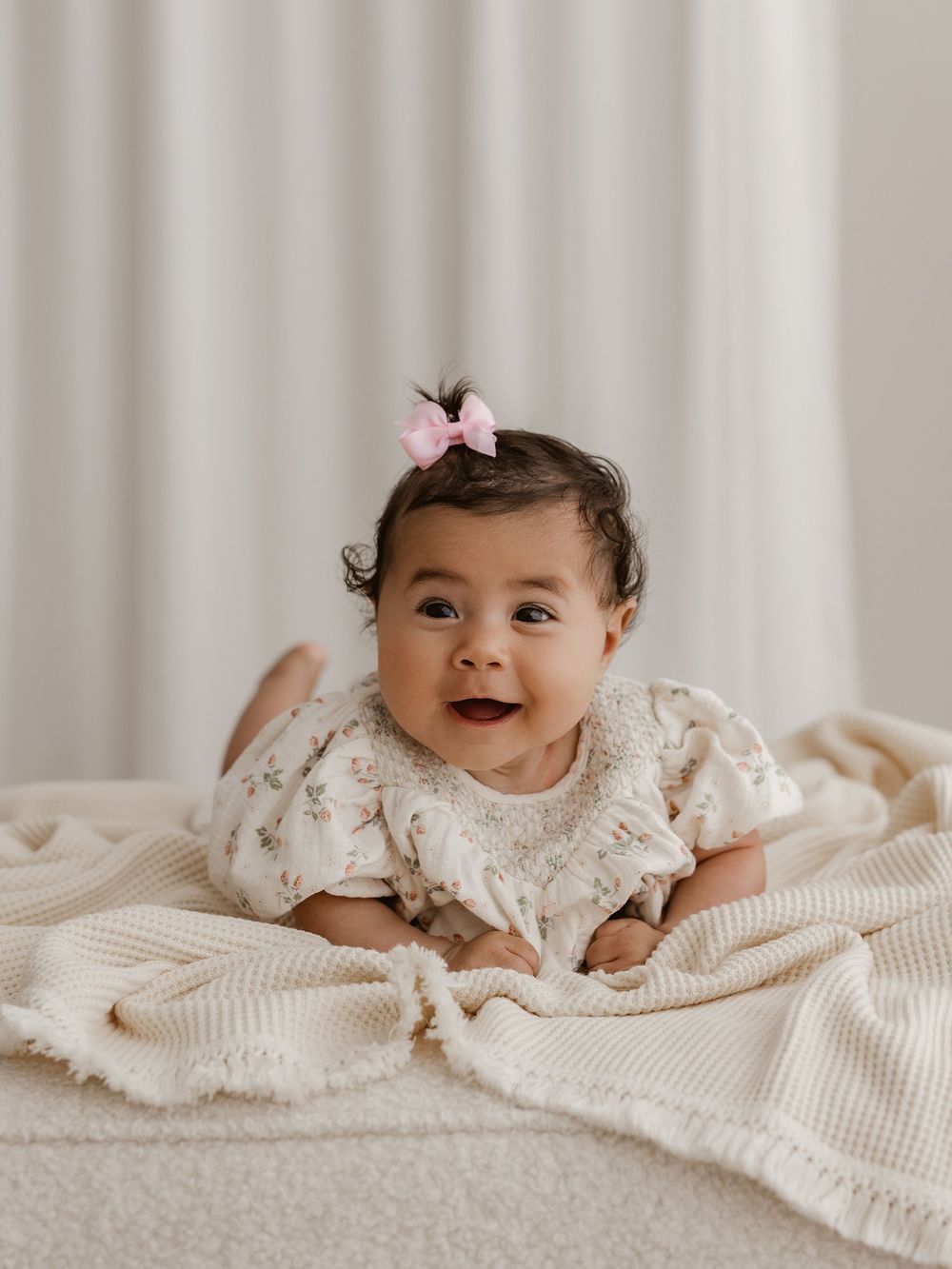 Baby in white lace dress lies on cream textured blanket while looking at camera in soft natural light.