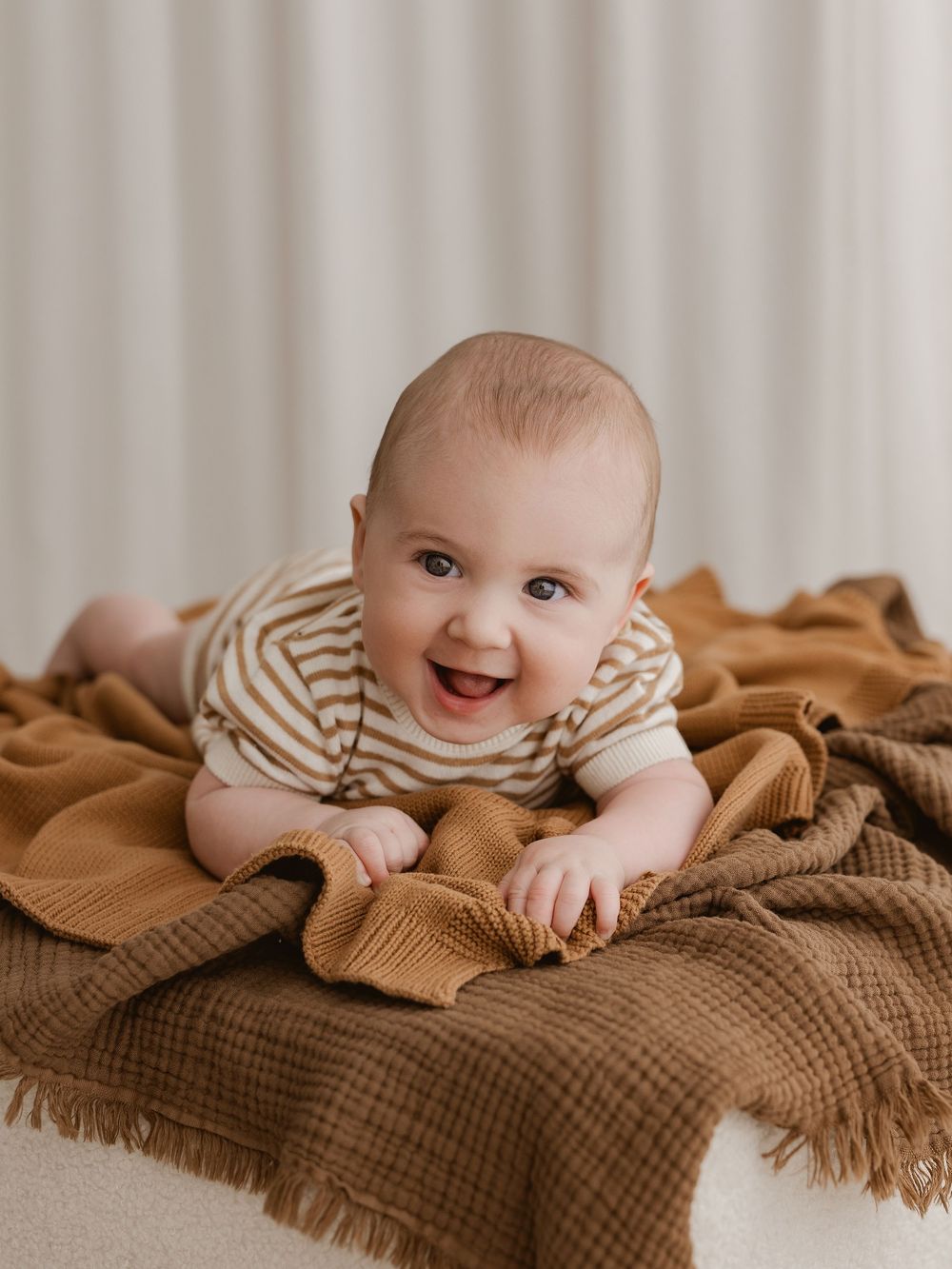 A baby lies on a brown knitted blanket, smiling and looking up at the camera in a series of progressive shots.