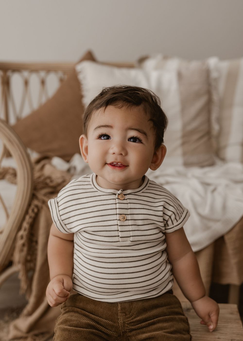 A baby wearing a striped shirt and brown pants smiles while lying on a beige couch.