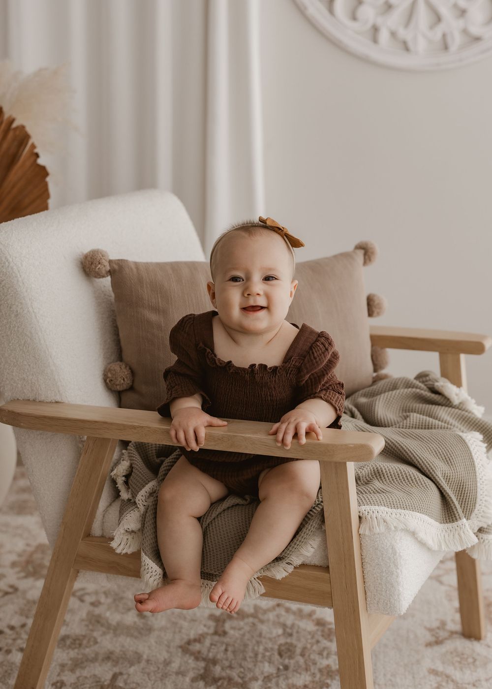 A baby in a brown outfit sits and plays on a wooden armchair with a beige cushion.