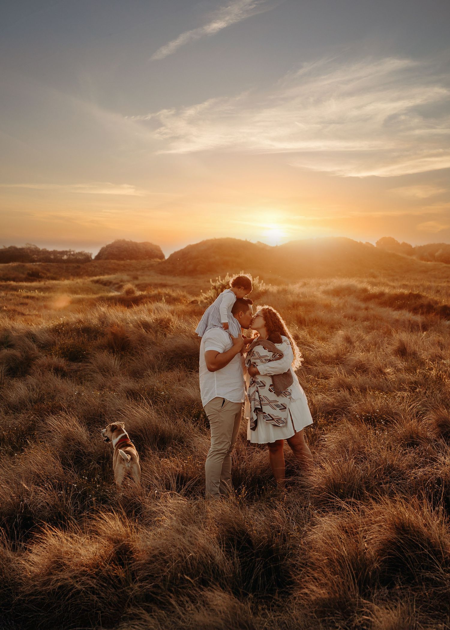 Family embracing during sunset photoshoot in golden grass field with dog.