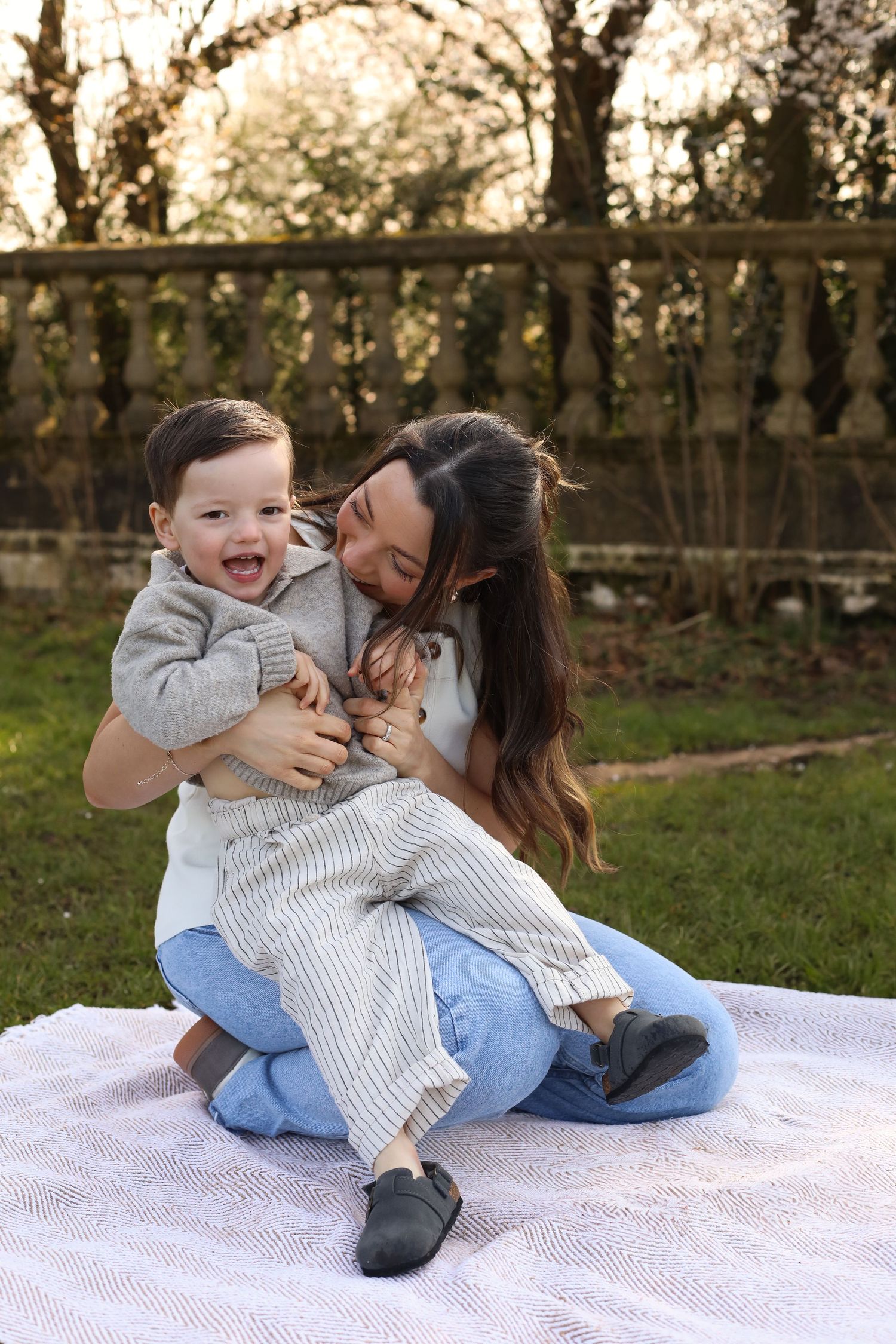 A parent and child share a joyful moment while sitting together on a white blanket in an outdoor garden setting.
