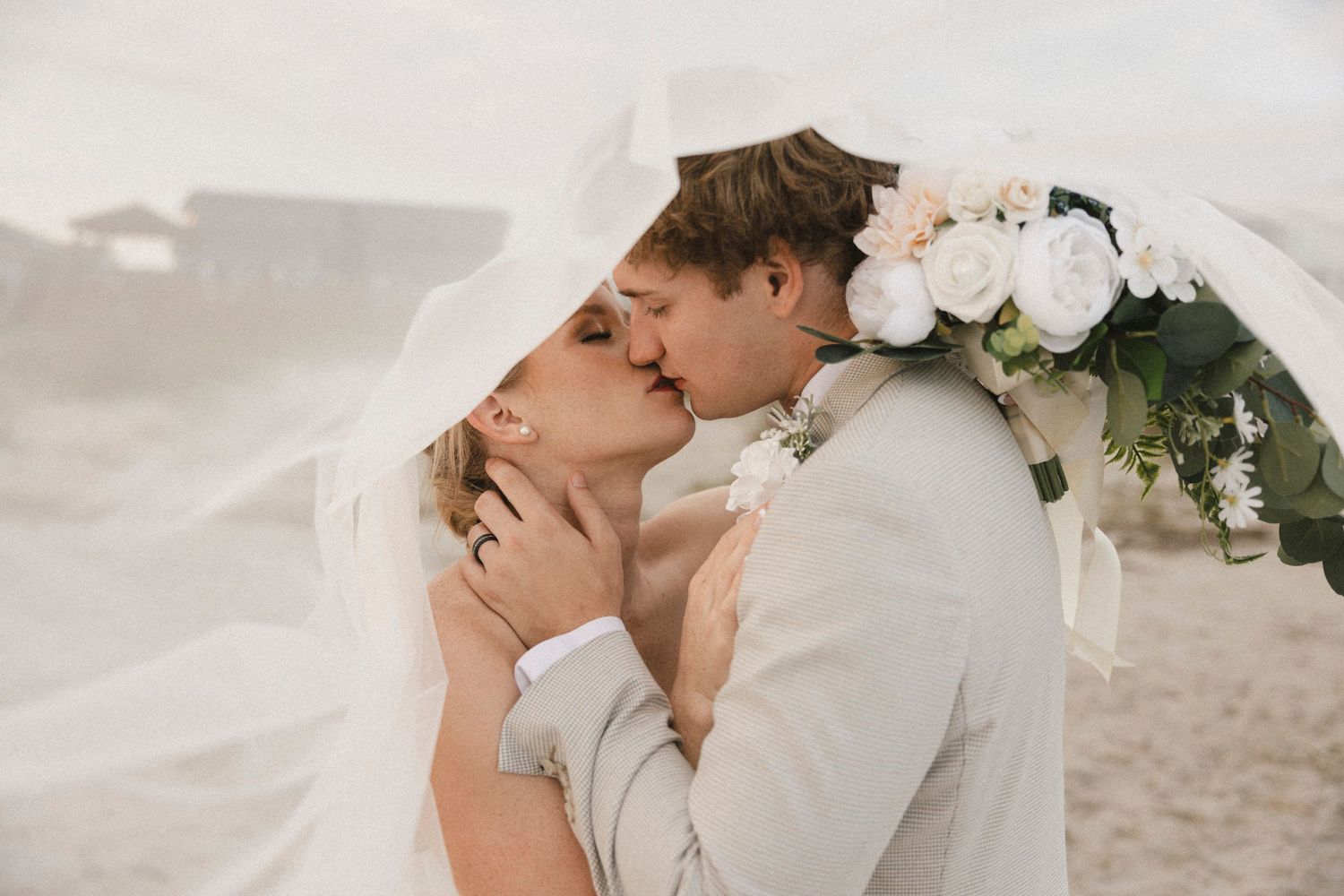 A romantic beach wedding moment with couple kissing under a veil adorned with white flowers.